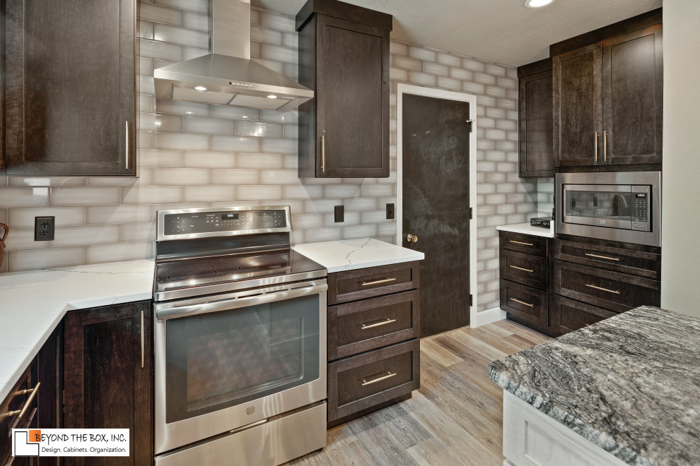 Mid-sized transitional l-shaped medium tone wood floor and brown floor kitchen photo with a farmhouse sink, shaker cabinets, black cabinets, granite countertops, gray backsplash, ceramic backsplash, stainless steel appliances, an island and white countertops