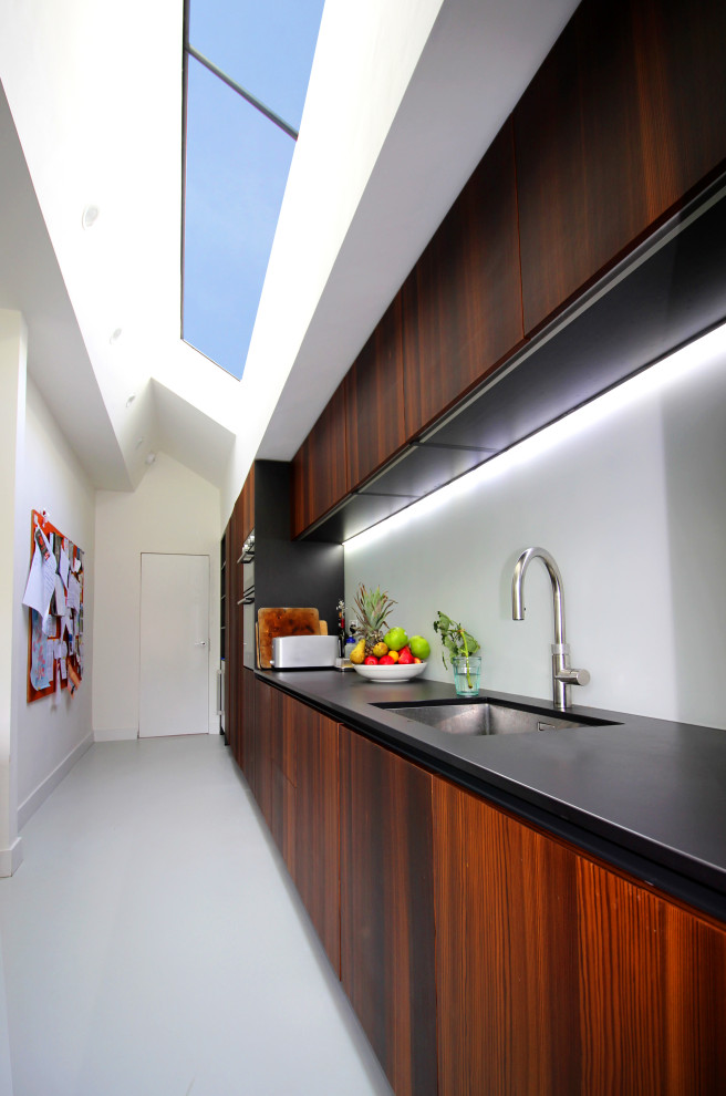 Photo of a mid-sized modern galley eat-in kitchen in London with a drop-in sink, flat-panel cabinets, dark wood cabinets, solid surface benchtops, green splashback, glass sheet splashback, black appliances, with island, grey floor and grey benchtop.
