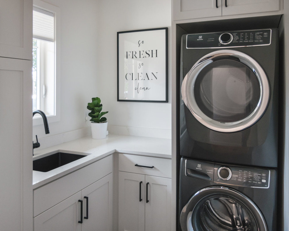 Utility room - small farmhouse l-shaped dark wood floor and brown floor utility room idea in Other with an undermount sink, shaker cabinets, white cabinets, quartz countertops, white backsplash, quartz backsplash, white walls, a stacked washer/dryer and white countertops