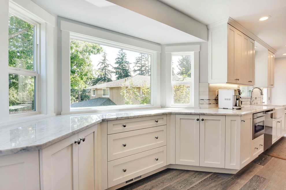 Mid-sized transitional galley eat-in kitchen in Seattle with a farmhouse sink, shaker cabinets, white cabinets, marble benchtops, white splashback, stone tile splashback, stainless steel appliances, medium hardwood floors and with island.