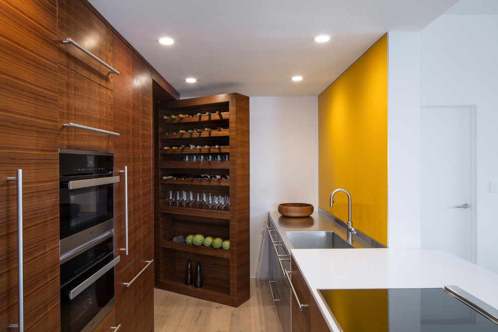 Modern galley kitchen in Los Angeles with an integrated sink, flat-panel cabinets, medium wood cabinets, stainless steel benchtops, yellow splashback, glass sheet splashback, stainless steel appliances, light hardwood floors, a peninsula, beige floor and grey benchtop.