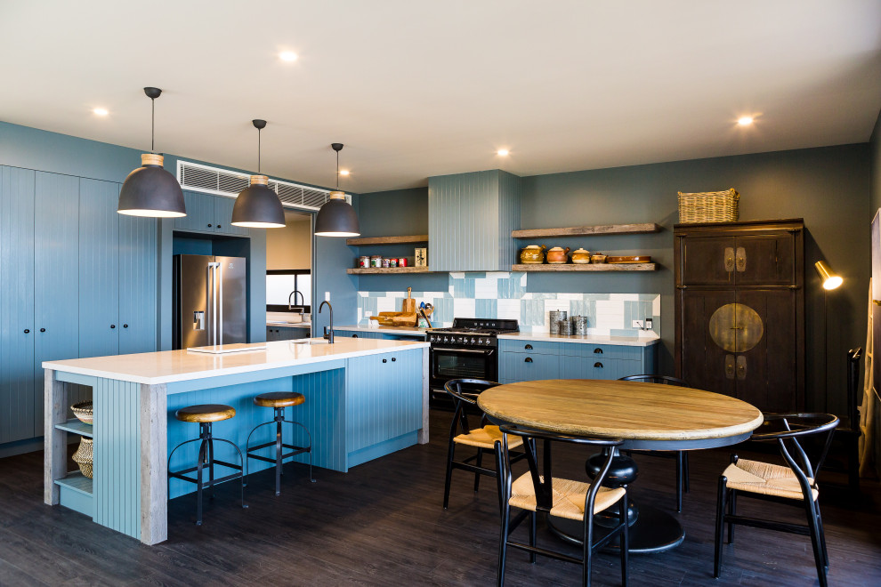 Photo of a beach style l-shaped kitchen in Wollongong with an undermount sink, flat-panel cabinets, blue cabinets, multi-coloured splashback, black appliances, dark hardwood floors, with island, brown floor and white benchtop.