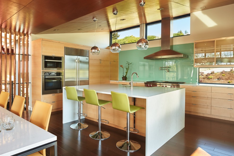 This is an example of a midcentury l-shaped kitchen in Seattle with flat-panel cabinets, light wood cabinets, green splashback, glass sheet splashback, stainless steel appliances, dark hardwood floors, with island, brown floor and white benchtop.
