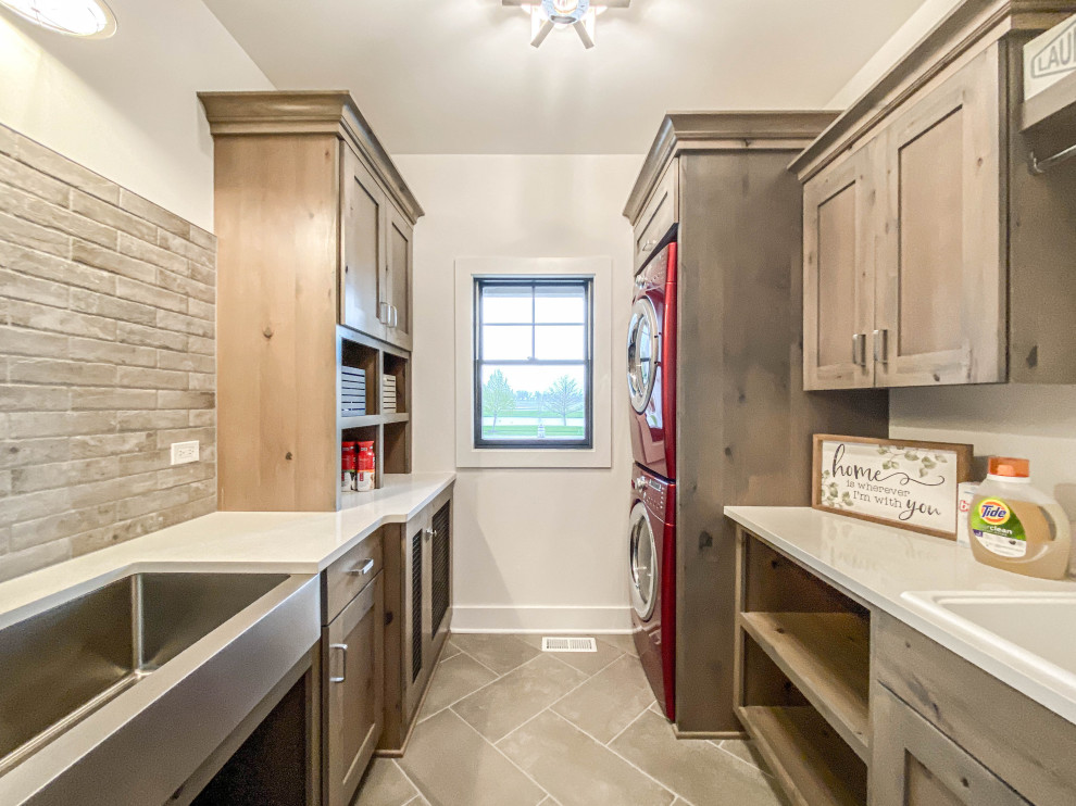 Mid-sized country dedicated laundry room in Chicago with a farmhouse sink, shaker cabinets, distressed cabinets, quartz benchtops, beige splashback, brick splashback, white walls, ceramic floors, a stacked washer and dryer, grey floor, white benchtop and brick walls.