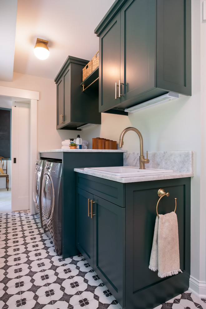 This is an example of a transitional l-shaped laundry room in New York with a drop-in sink, shaker cabinets, green cabinets, granite benchtops, grey walls, ceramic floors, a side-by-side washer and dryer, white floor and white benchtop.