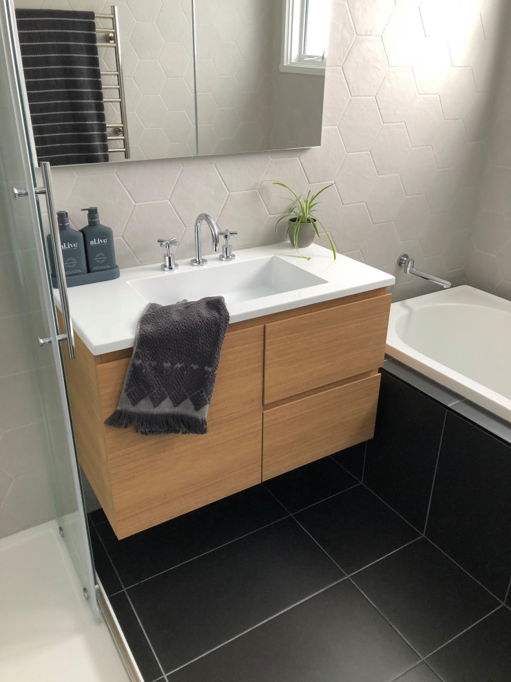Inspiration for a small contemporary master bathroom with flat-panel cabinets, light wood cabinets, a drop-in tub, a corner shower, gray tile, ceramic tile, grey walls, ceramic floors, an integrated sink, solid surface benchtops, black floor, a sliding shower screen, white benchtops, a single vanity and a floating vanity.