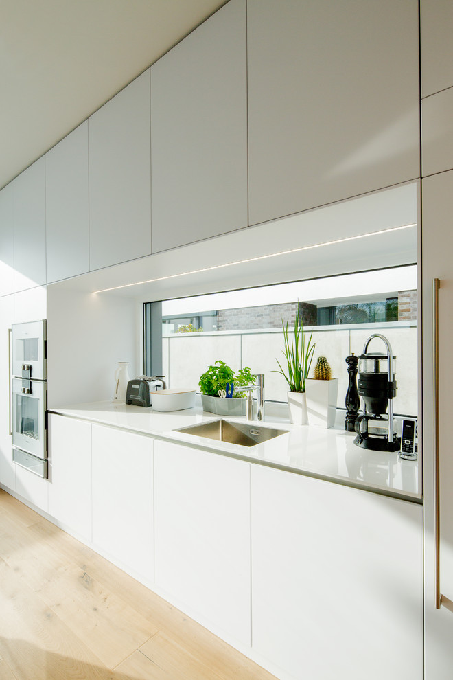 Photo of a large modern galley open plan kitchen in Dusseldorf with an undermount sink, flat-panel cabinets, white cabinets, solid surface benchtops, white splashback, glass sheet splashback, white appliances, medium hardwood floors, with island, brown floor and white benchtop.
