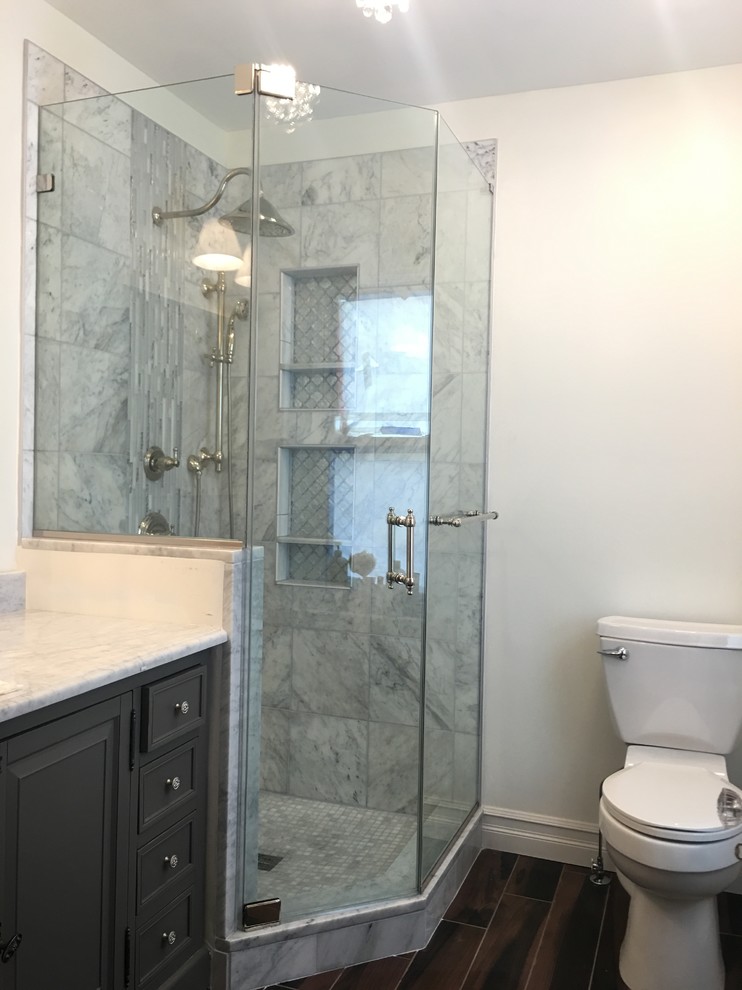 This is an example of a mid-sized traditional master bathroom in Newark with recessed-panel cabinets, grey cabinets, an alcove shower, a one-piece toilet, gray tile, white tile, porcelain tile, white walls, dark hardwood floors and marble benchtops.
