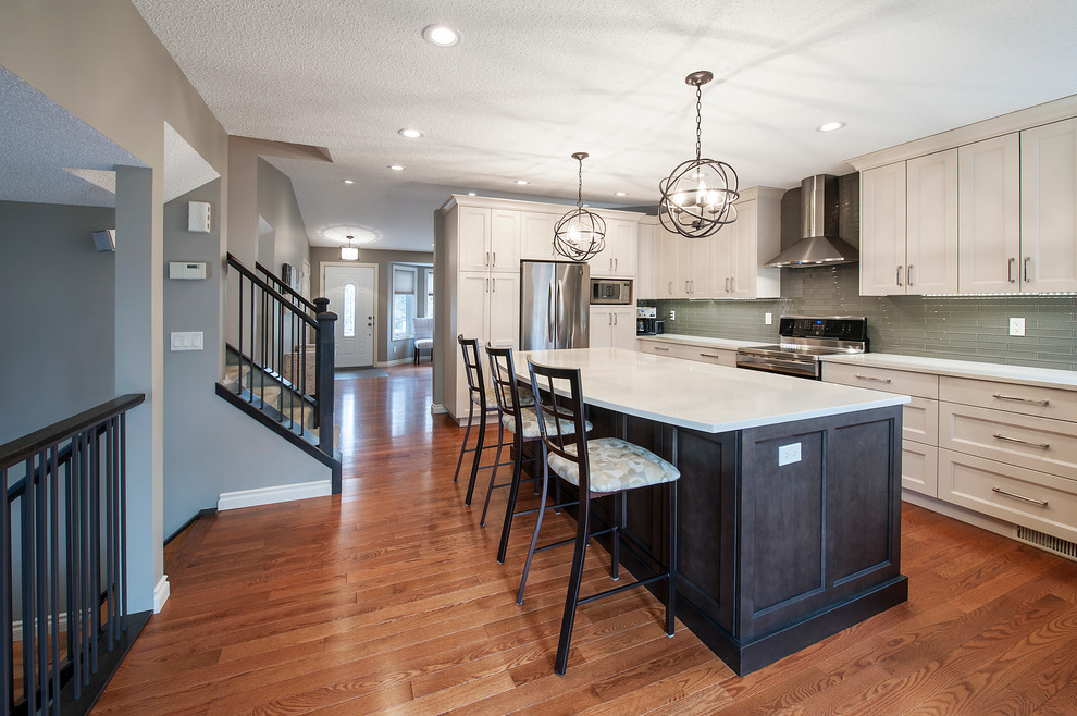 Large transitional l-shaped dark wood floor eat-in kitchen photo in Calgary with an undermount sink, recessed-panel cabinets, white cabinets, solid surface countertops, green backsplash, glass tile backsplash, stainless steel appliances and an island