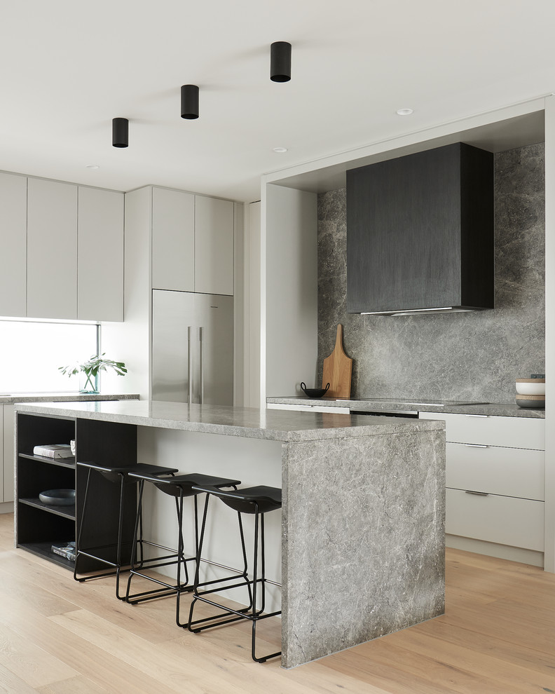 Modern l-shaped kitchen in Melbourne with flat-panel cabinets, white cabinets, grey splashback, stone slab splashback, stainless steel appliances, light hardwood floors, with island, grey benchtop and beige floor.