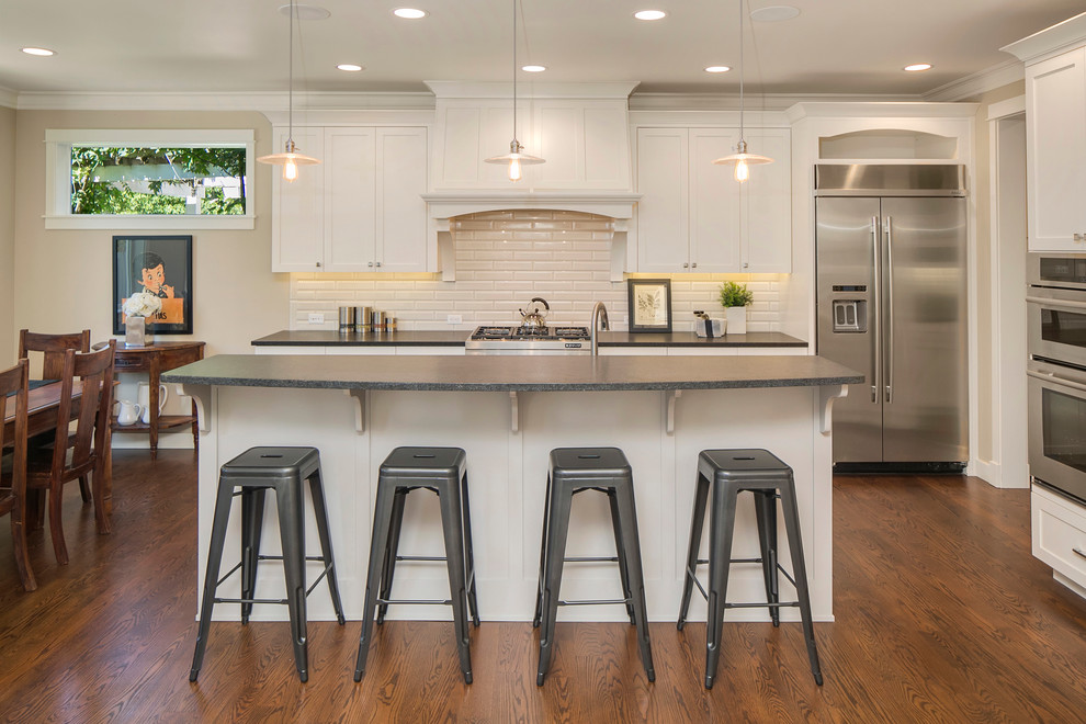 Eat-in kitchen - traditional galley medium tone wood floor eat-in kitchen idea in Seattle with shaker cabinets, white cabinets, an island, a farmhouse sink, granite countertops, gray backsplash, subway tile backsplash and stainless steel appliances