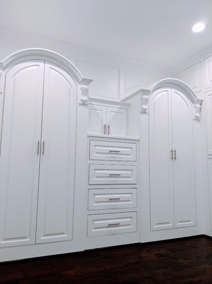 This is an example of a large modern gender-neutral walk-in wardrobe in Houston with raised-panel cabinets, white cabinets, dark hardwood floors, brown floor and vaulted.