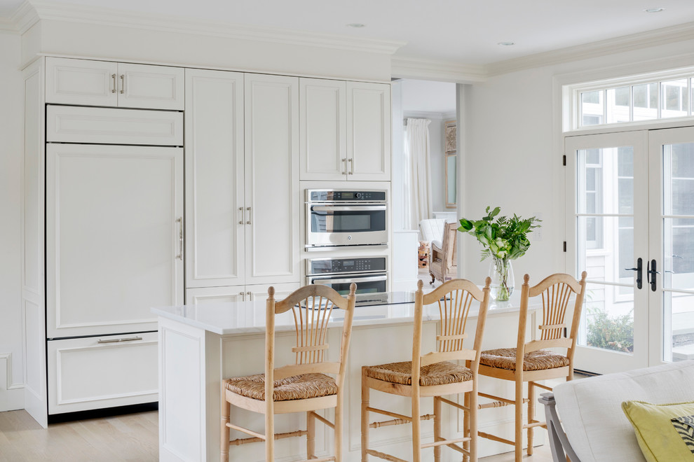 Large transitional l-shaped open plan kitchen in Providence with white cabinets, white splashback, panelled appliances, light hardwood floors, with island, an undermount sink, recessed-panel cabinets and marble benchtops.