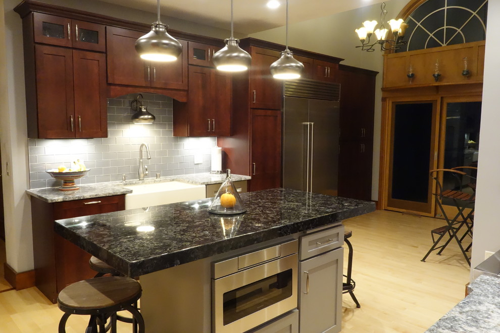 Mid-sized light wood floor eat-in kitchen photo with a farmhouse sink, recessed-panel cabinets, dark wood cabinets, granite countertops, gray backsplash, glass tile backsplash, stainless steel appliances and an island