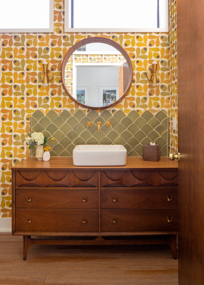 Mid-sized midcentury powder room in Detroit with furniture-like cabinets, brown cabinets, a one-piece toilet, multi-coloured tile, ceramic tile, multi-coloured walls, medium hardwood floors, a vessel sink, wood benchtops, brown floor, brown benchtops, a freestanding vanity, vaulted and wallpaper.
