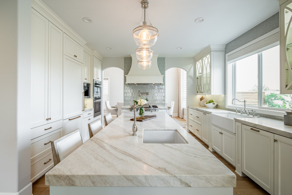 Design ideas for a mid-sized traditional kitchen in Orange County with a farmhouse sink, recessed-panel cabinets, blue cabinets, quartzite benchtops, blue splashback, ceramic splashback, panelled appliances, light hardwood floors, with island and beige benchtop.