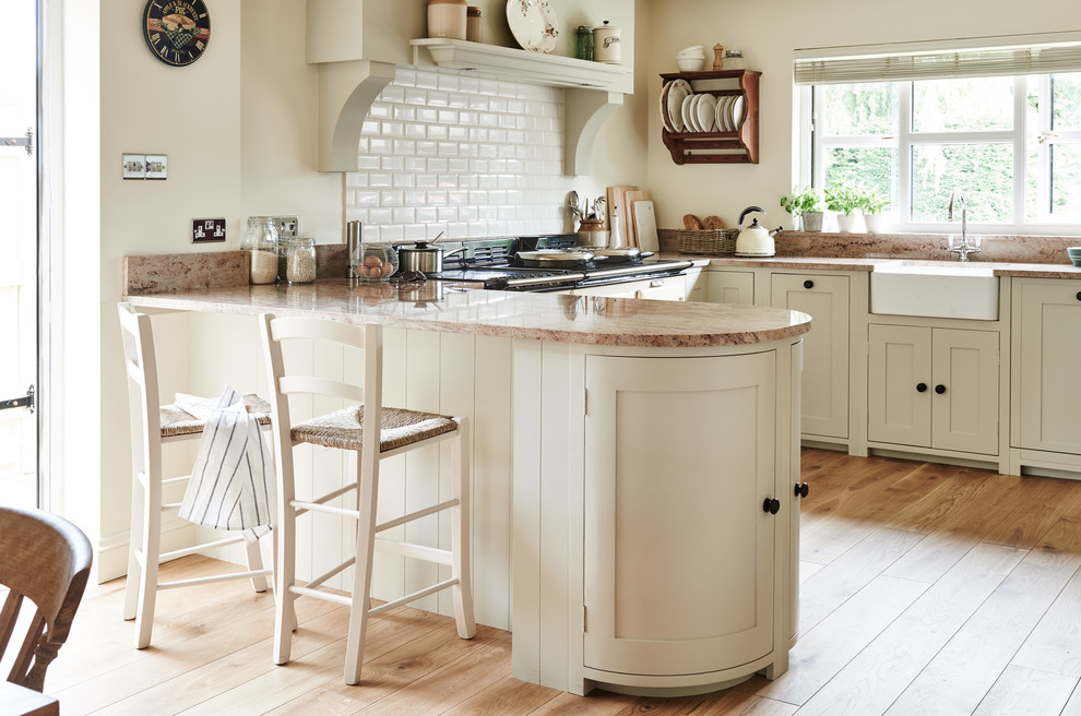 Country u-shaped kitchen in Wiltshire with a farmhouse sink, shaker cabinets, beige cabinets, white splashback, subway tile splashback, light hardwood floors, a peninsula, beige floor and beige benchtop.