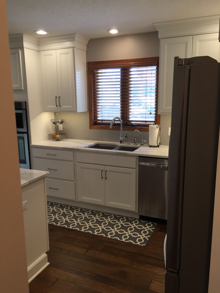 Example of a large transitional l-shaped dark wood floor home design design with an undermount sink, flat-panel cabinets, white cabinets, quartz countertops, white backsplash, ceramic backsplash, stainless steel appliances and an island