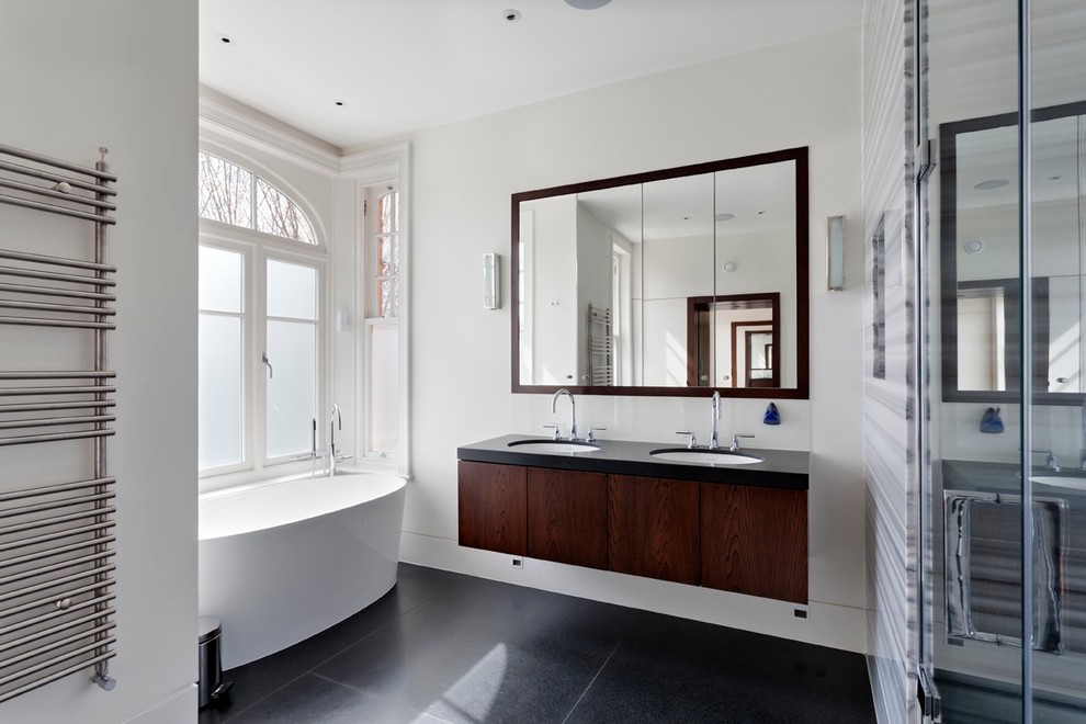 Design ideas for a large contemporary master bathroom in London with flat-panel cabinets, dark wood cabinets, a freestanding tub, an open shower, a wall-mount toilet, gray tile, marble, grey walls, slate floors, a drop-in sink, limestone benchtops, black floor, a hinged shower door and black benchtops.