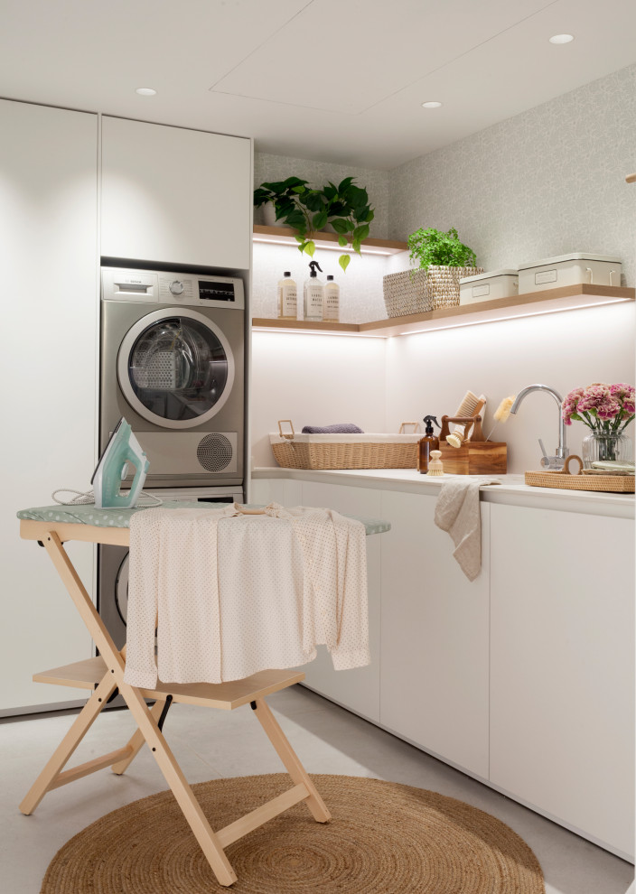 Large l-shaped dedicated laundry room in Valencia with an undermount sink, flat-panel cabinets, white cabinets, ceramic floors, a stacked washer and dryer, beige floor, white benchtop and wallpaper.
