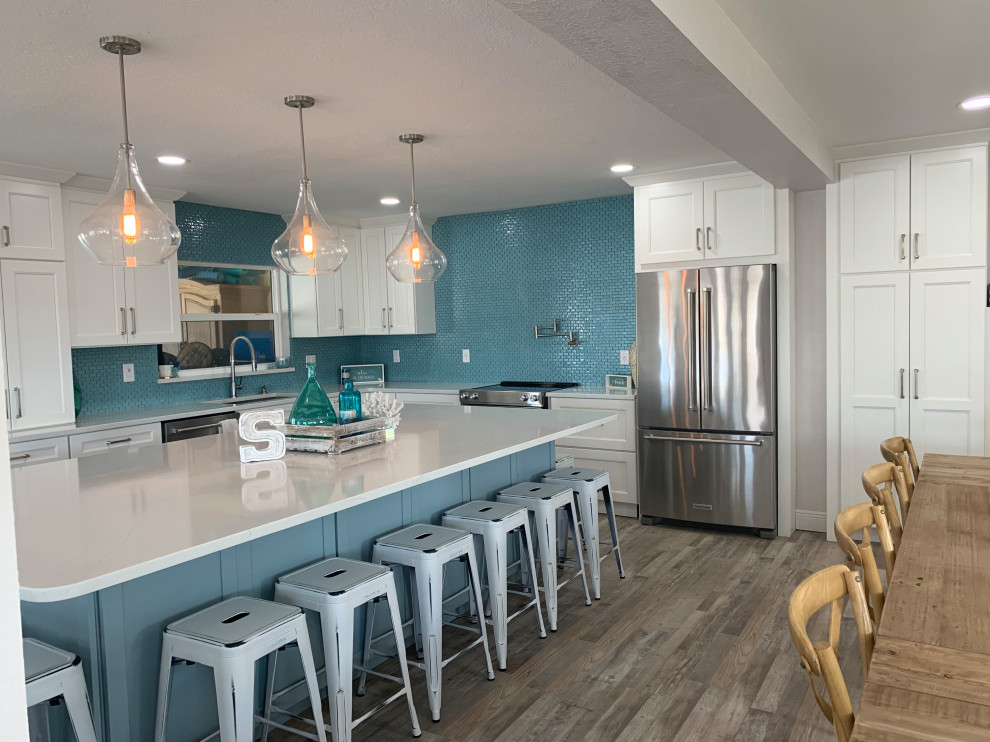 Photo of a large beach style l-shaped eat-in kitchen in Tampa with an undermount sink, shaker cabinets, blue cabinets, quartz benchtops, blue splashback, glass tile splashback, stainless steel appliances, vinyl floors, with island, brown floor and white benchtop.