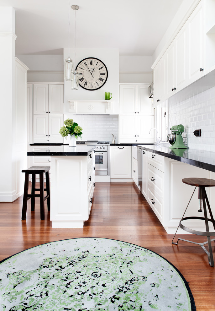 Photo of a transitional l-shaped kitchen in Melbourne with a farmhouse sink, recessed-panel cabinets, white splashback, subway tile splashback, stainless steel appliances, medium hardwood floors, with island and black benchtop.