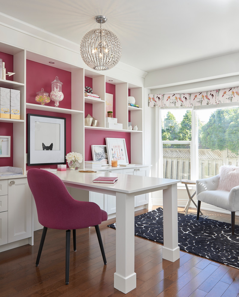 Photo of a transitional craft room in Toronto with pink walls, medium hardwood floors, no fireplace, a built-in desk and brown floor.