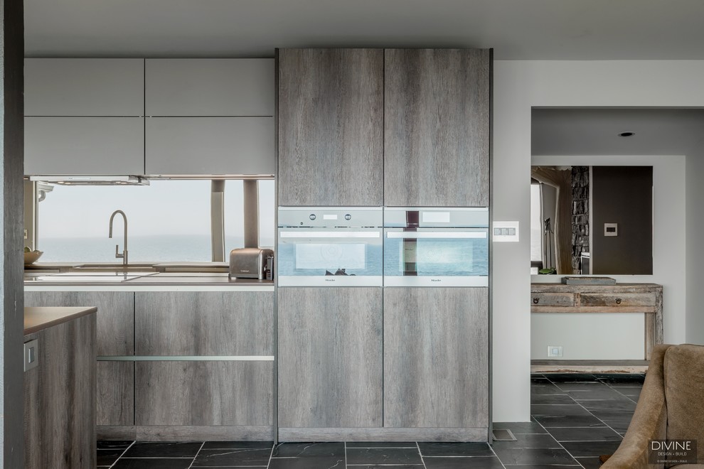 This is an example of a large eclectic single-wall open plan kitchen in Boston with a farmhouse sink, flat-panel cabinets, dark wood cabinets, granite benchtops, metallic splashback, glass sheet splashback, stainless steel appliances, slate floors, with island, grey floor and brown benchtop.