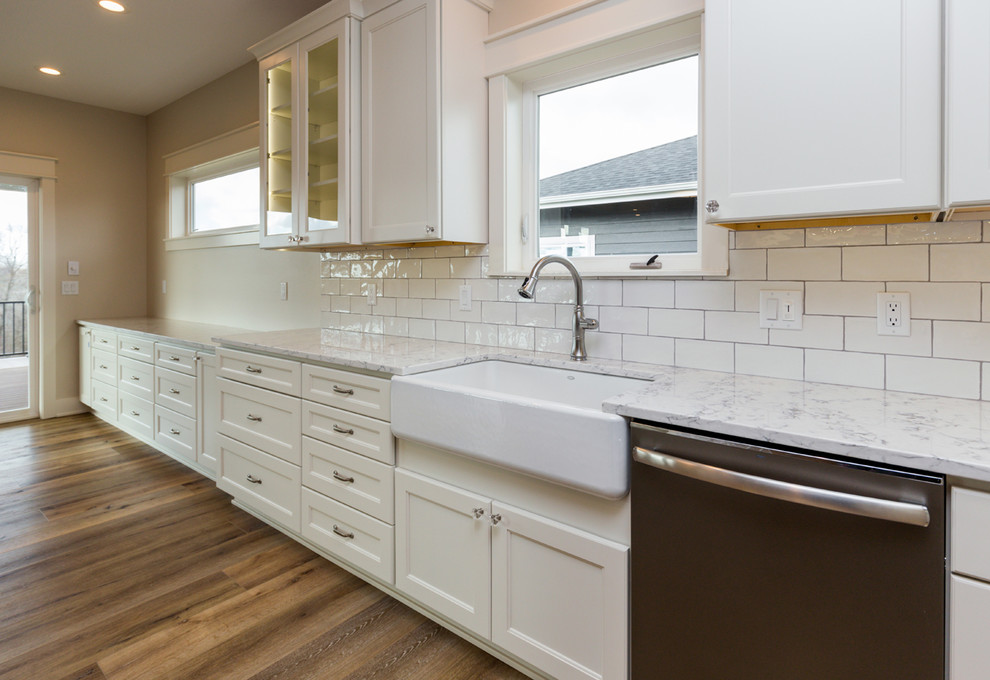 Photo of a country l-shaped eat-in kitchen in Other with a farmhouse sink, shaker cabinets, white cabinets, quartz benchtops, white splashback, ceramic splashback, stainless steel appliances, vinyl floors, with island and brown floor.