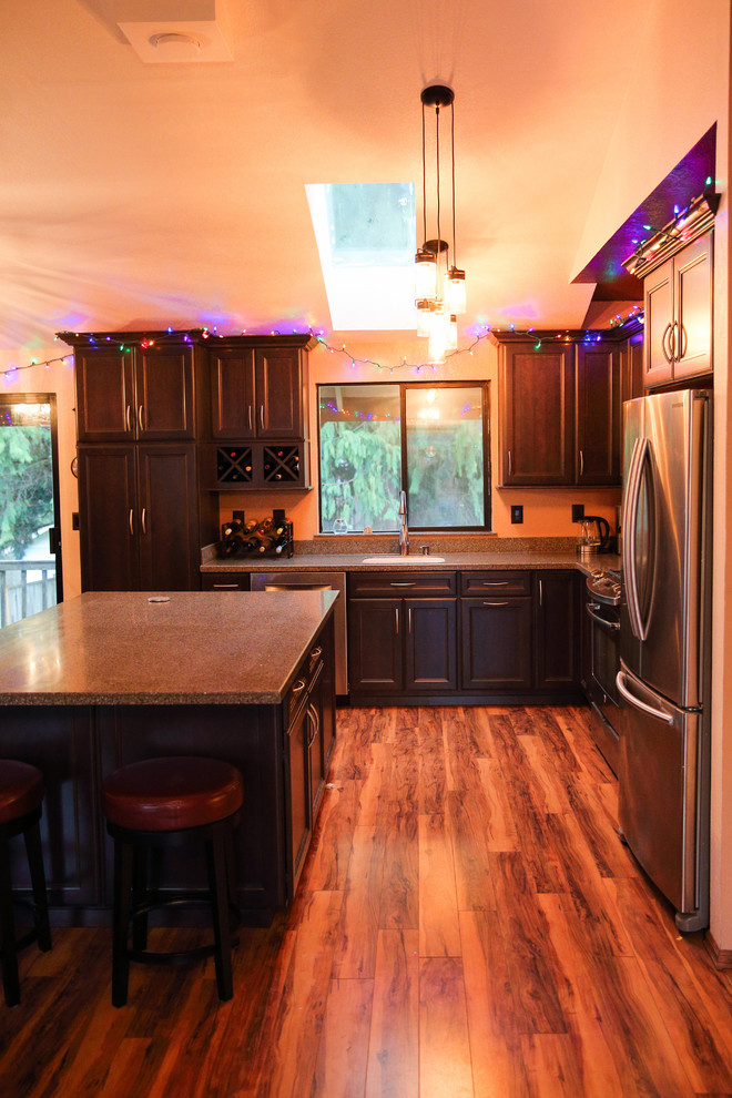 Example of a mid-sized transitional l-shaped medium tone wood floor open concept kitchen design in Seattle with an integrated sink, flat-panel cabinets, dark wood cabinets, solid surface countertops, stainless steel appliances and an island