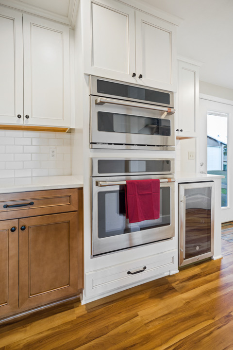 Inspiration for a mid-sized transitional l-shaped medium tone wood floor and brown floor eat-in kitchen remodel with a farmhouse sink, shaker cabinets, white cabinets, quartz countertops, white backsplash, subway tile backsplash, stainless steel appliances, an island and white countertops