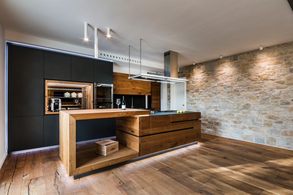 Large contemporary single-wall open plan kitchen in Rome with a drop-in sink, black cabinets, wood benchtops, black splashback, slate splashback, medium hardwood floors, with island and flat-panel cabinets.