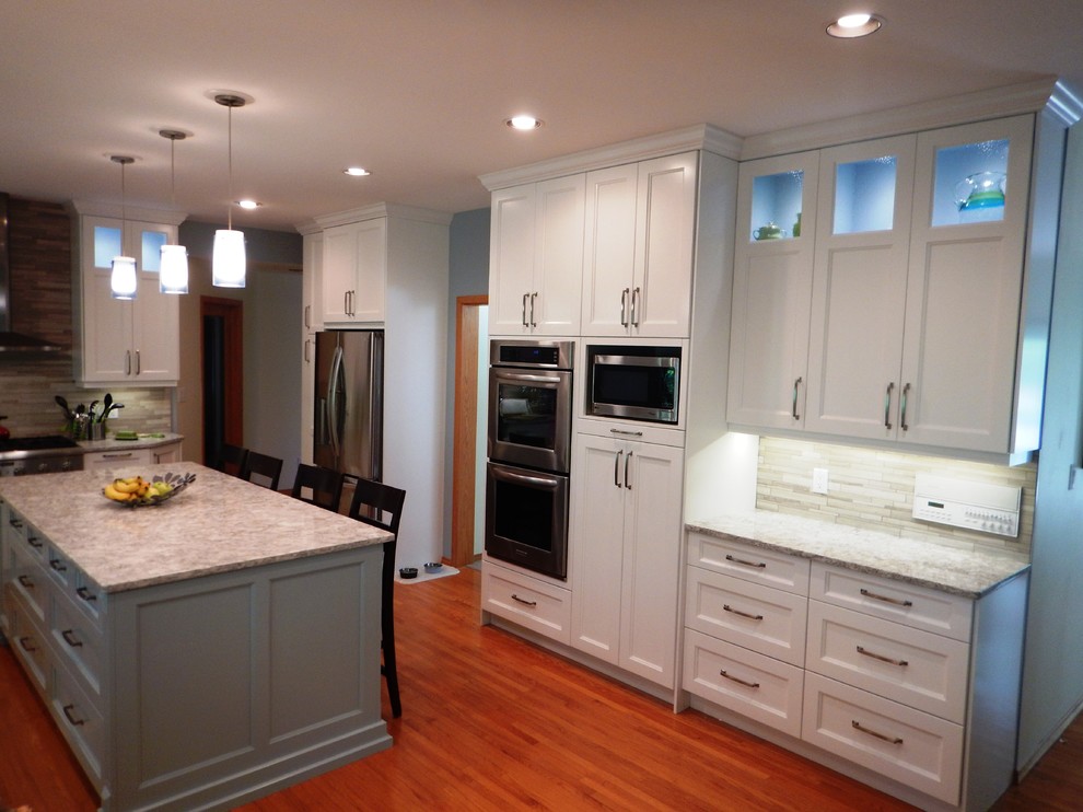 Example of a large transitional l-shaped light wood floor eat-in kitchen design with an undermount sink, recessed-panel cabinets, white cabinets, quartz countertops, gray backsplash, stone tile backsplash, stainless steel appliances and an island