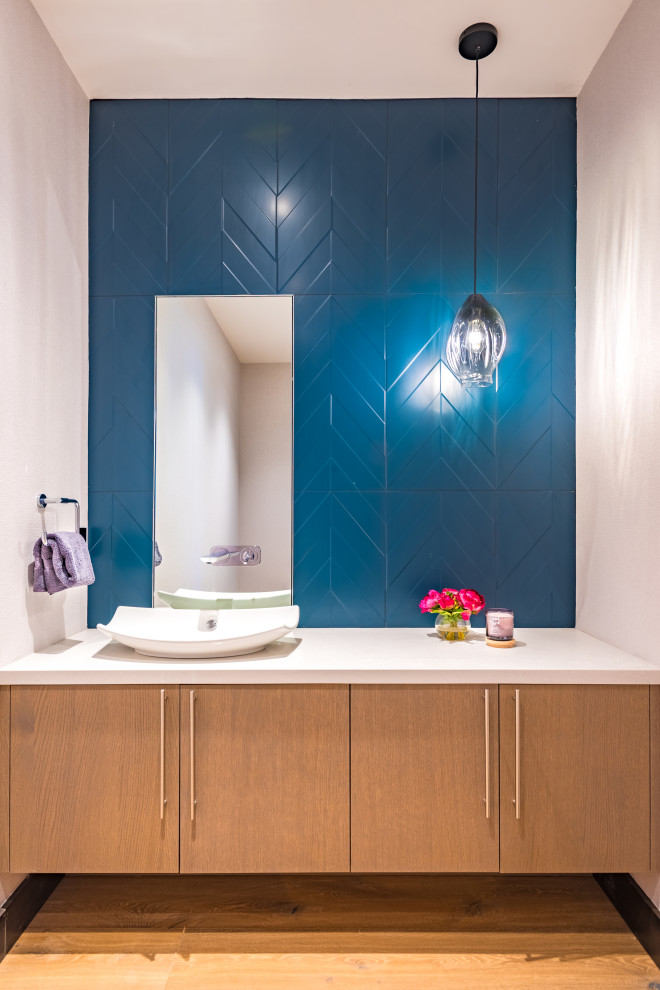 This is an example of a mid-sized modern powder room with flat-panel cabinets, medium wood cabinets, a two-piece toilet, blue tile, porcelain tile, beige walls, medium hardwood floors, a vessel sink, engineered quartz benchtops, brown floor, grey benchtops, a floating vanity and wallpaper.