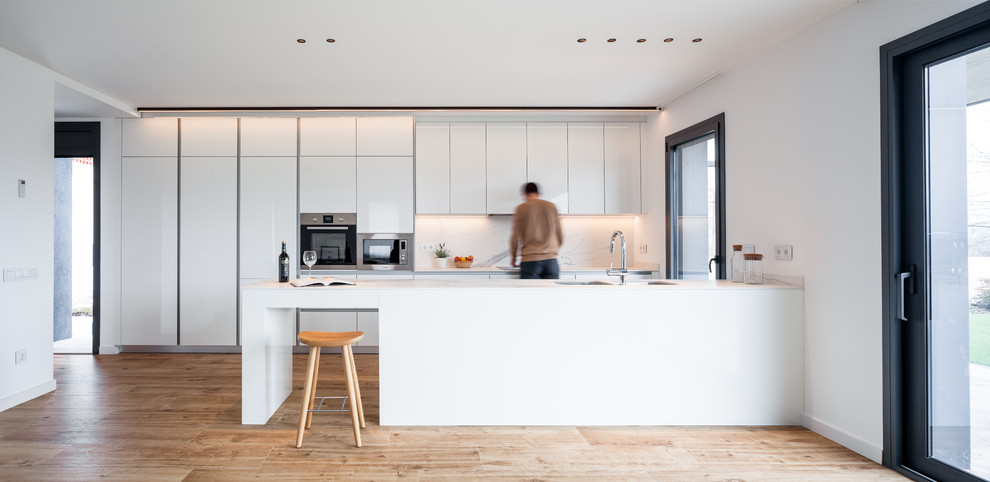 This is an example of a contemporary galley kitchen in Other with a double-bowl sink, flat-panel cabinets, white cabinets, white splashback, light hardwood floors, a peninsula, brown floor and white benchtop.