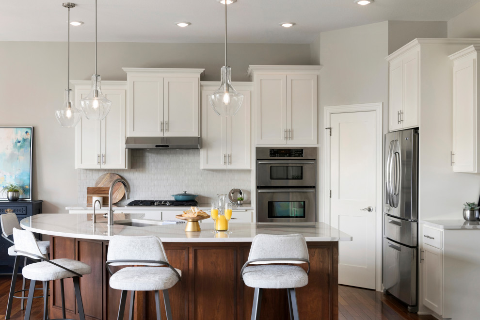 Photo of a large beach style u-shaped eat-in kitchen in Minneapolis with a drop-in sink, recessed-panel cabinets, white cabinets, quartzite benchtops, white splashback, ceramic splashback, stainless steel appliances, medium hardwood floors, with island, brown floor and white benchtop.