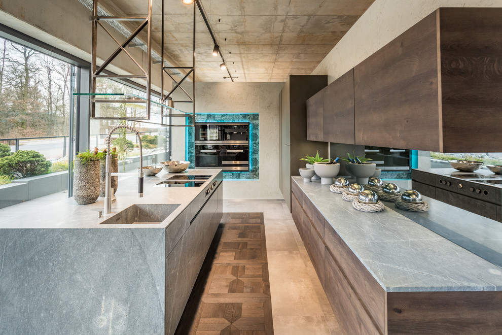This is an example of a large industrial kitchen in Frankfurt with an integrated sink, flat-panel cabinets, dark wood cabinets, granite benchtops, mirror splashback, with island, grey benchtop and black appliances.