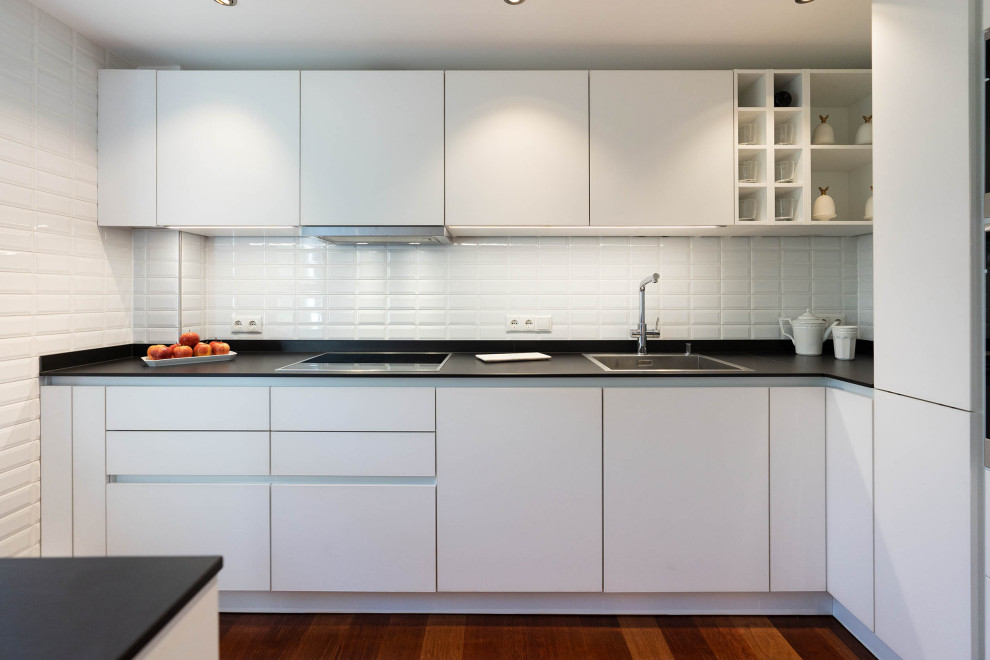 Example of a small trendy galley medium tone wood floor and brown floor enclosed kitchen design in Berlin with a drop-in sink, flat-panel cabinets, white cabinets, quartzite countertops, white backsplash, subway tile backsplash, black appliances and black countertops