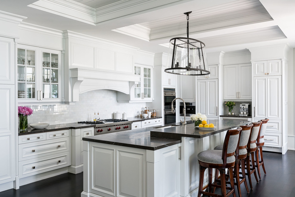 This is an example of a traditional l-shaped kitchen with an undermount sink, raised-panel cabinets, white cabinets, white splashback, subway tile splashback, stainless steel appliances, dark hardwood floors, with island, black floor and black benchtop.