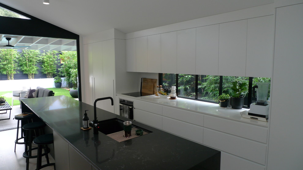 Design ideas for a mid-sized modern galley open plan kitchen in Sydney with an undermount sink, flat-panel cabinets, black cabinets, solid surface benchtops, white splashback, ceramic splashback, black appliances, painted wood floors, with island, white floor and black benchtop.