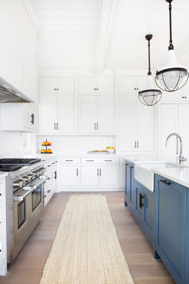 This is an example of a large beach style u-shaped eat-in kitchen in New York with a farmhouse sink, shaker cabinets, white cabinets, quartz benchtops, white splashback, timber splashback, stainless steel appliances, light hardwood floors, with island, beige floor and white benchtop.