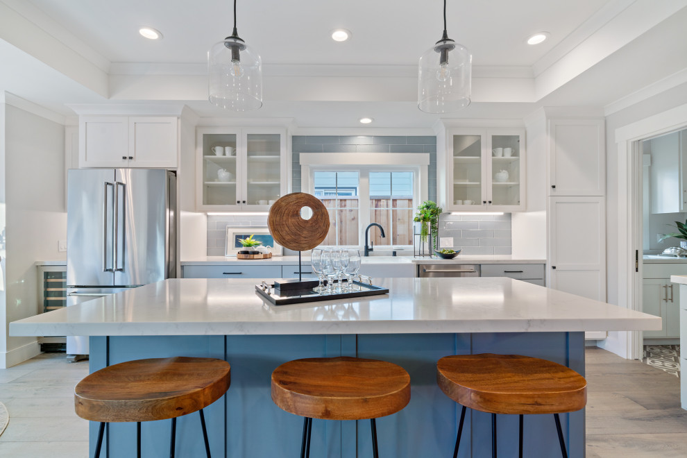 Mid-sized arts and crafts l-shaped open plan kitchen in San Francisco with a farmhouse sink, shaker cabinets, white cabinets, quartz benchtops, blue splashback, glass tile splashback, stainless steel appliances, light hardwood floors, with island, grey floor and white benchtop.