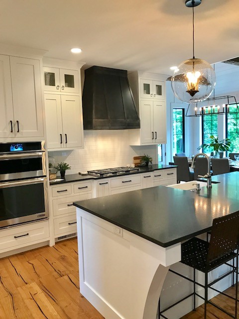 This is an example of a large country l-shaped eat-in kitchen in Grand Rapids with white cabinets, with island, a farmhouse sink, shaker cabinets, soapstone benchtops, white splashback, subway tile splashback, panelled appliances, light hardwood floors, brown floor and black benchtop.