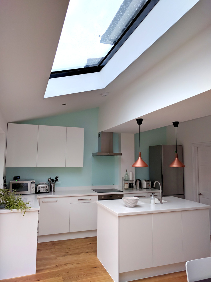 Photo of a mid-sized contemporary l-shaped open plan kitchen in London with an integrated sink, flat-panel cabinets, white cabinets, solid surface benchtops, blue splashback, glass sheet splashback, white appliances, light hardwood floors, with island and beige floor.