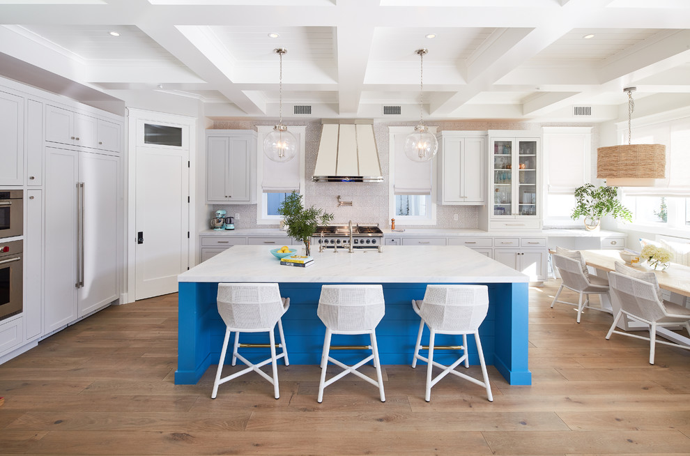 This is an example of a beach style l-shaped eat-in kitchen in Los Angeles with a farmhouse sink, shaker cabinets, blue cabinets, beige splashback, panelled appliances, medium hardwood floors, with island, brown floor and white benchtop.