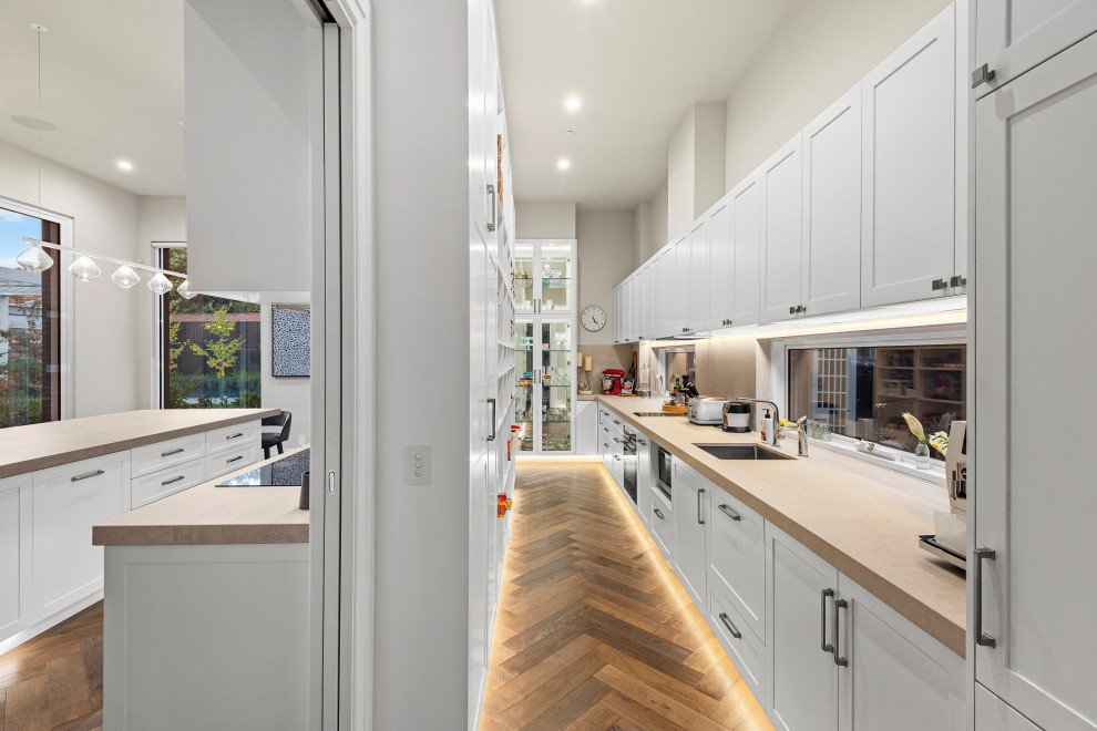 Photo of a large contemporary galley kitchen pantry in Melbourne with a single-bowl sink, recessed-panel cabinets, white cabinets, window splashback, stainless steel appliances, medium hardwood floors, no island, brown floor, beige benchtop and granite benchtops.