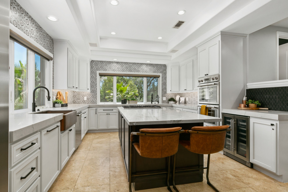 Large transitional u-shaped open plan kitchen in Orange County with a farmhouse sink, recessed-panel cabinets, white cabinets, marble benchtops, multi-coloured splashback, porcelain splashback, stainless steel appliances, limestone floors, with island, beige floor, grey benchtop and vaulted.