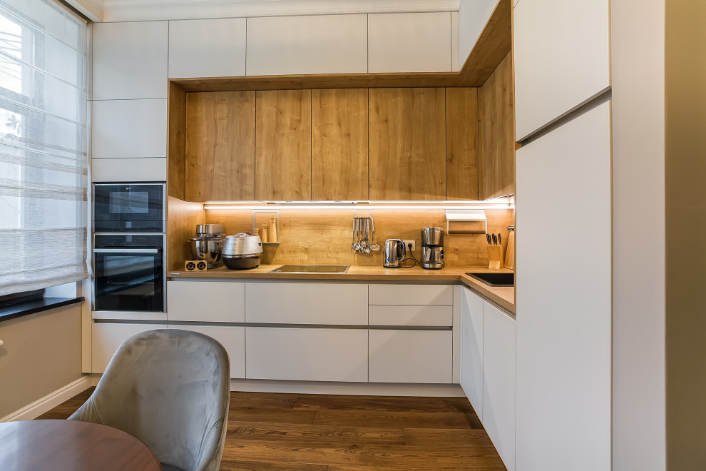 This is an example of a large scandi l-shaped kitchen/diner in Other with a single-bowl sink, glass-front cabinets, white cabinets, laminate countertops, brown splashback, black appliances, painted wood flooring, no island, brown floors and brown worktops.