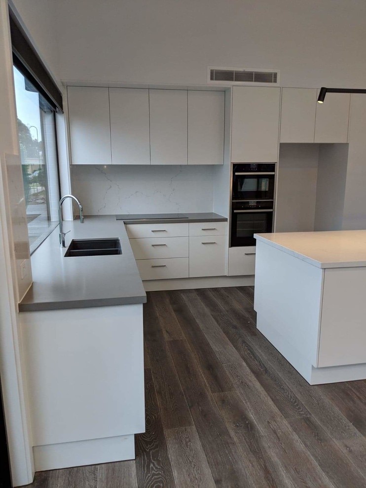Design ideas for a mid-sized modern l-shaped eat-in kitchen in Melbourne with an undermount sink, flat-panel cabinets, white cabinets, quartz benchtops, white splashback, stone slab splashback, black appliances, laminate floors, with island and grey benchtop.