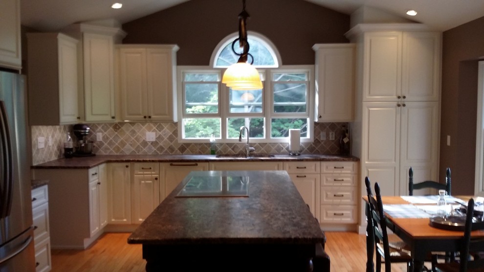 Mid-sized transitional l-shaped medium tone wood floor and brown floor kitchen photo with an undermount sink, raised-panel cabinets, white cabinets, brown backsplash, ceramic backsplash, stainless steel appliances and an island